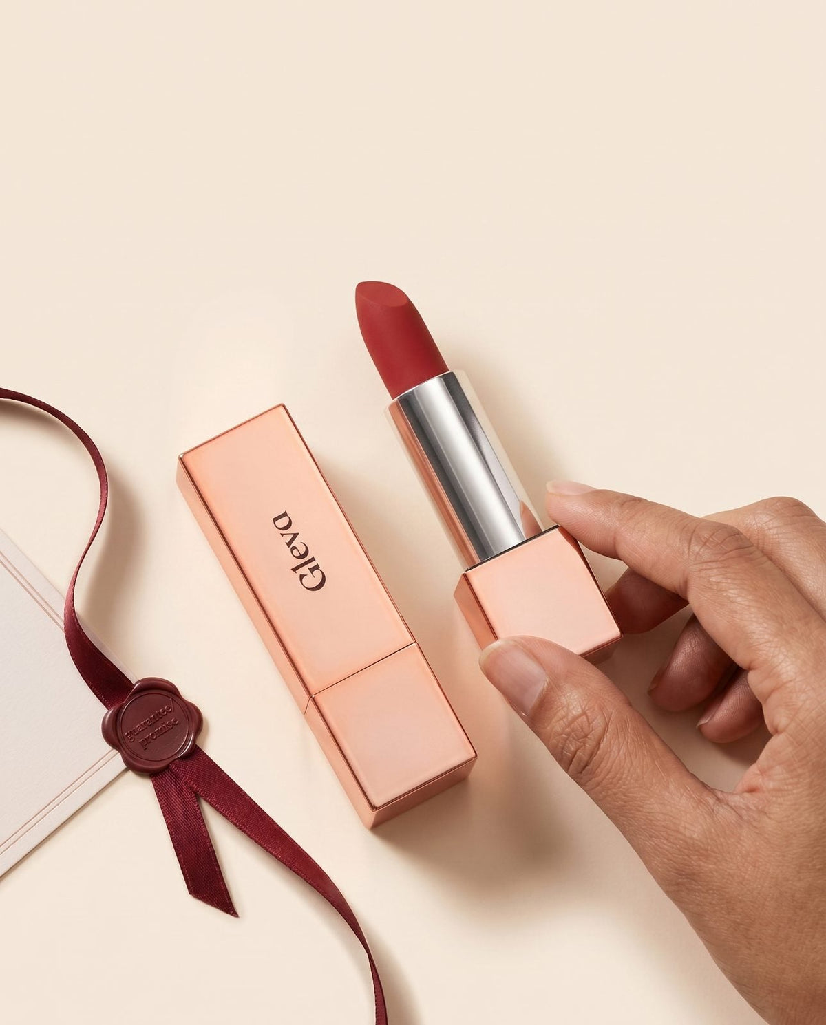 Hand holding a Creamy Matte Lipstick beside its rose-gold case and an oxblood ribbon