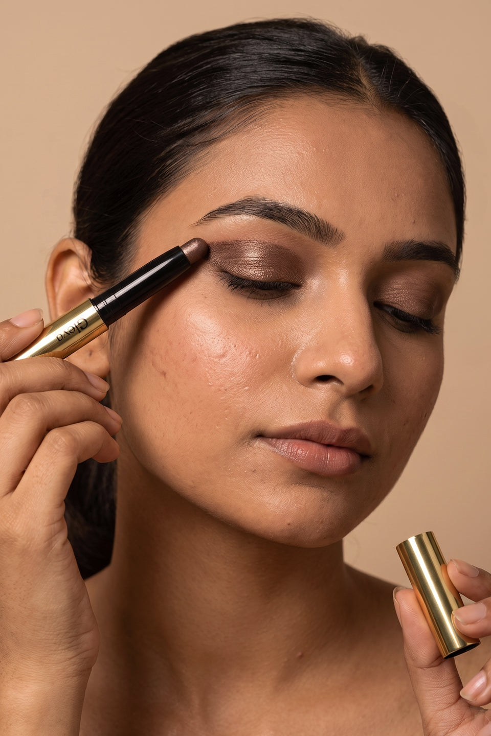 A woman swiping a bronze Gleva Eyeshadow Stick across her closed eyelid, the gold cap in her other hand