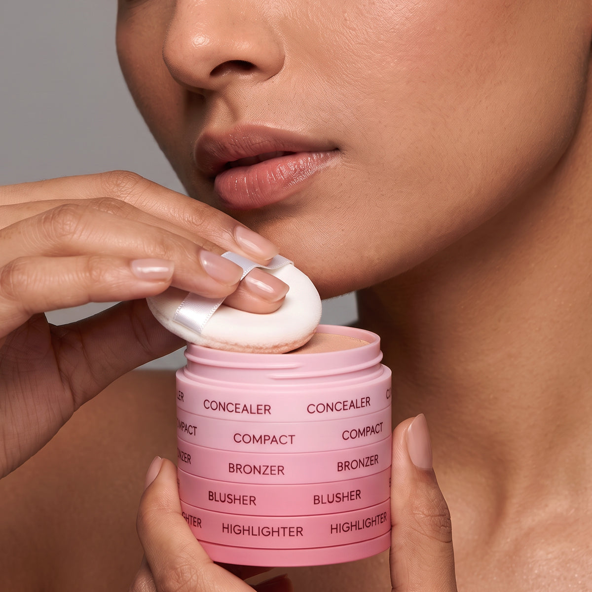 A woman pressing the white puff into the open top pan of the layer-labelled Makeup Stack, held at her chin